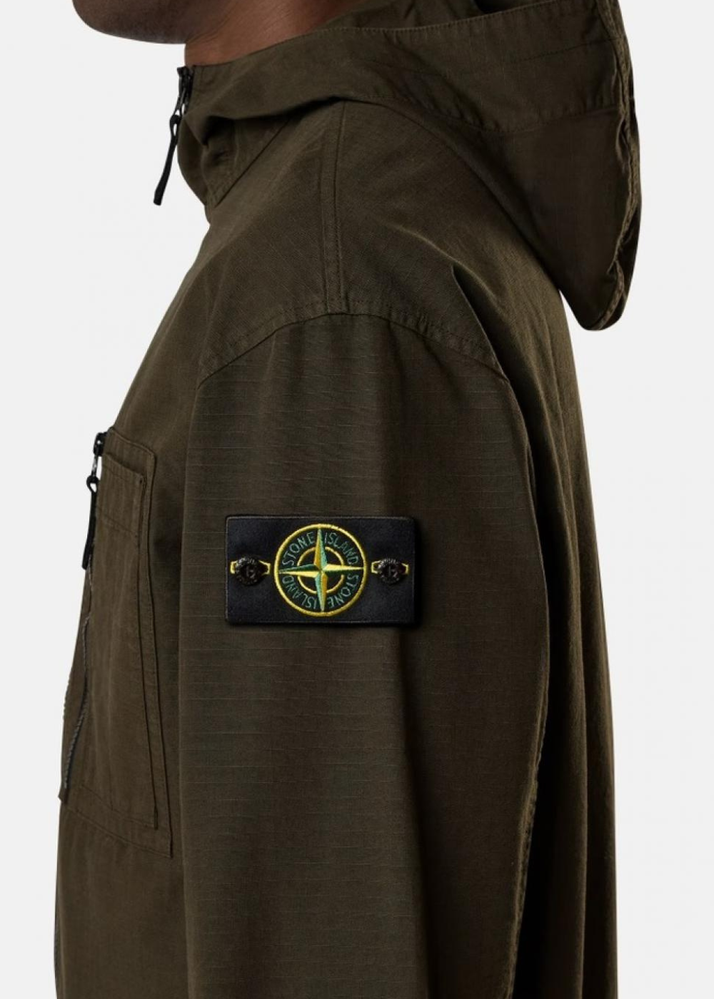 Zoom de profil sur la manche gauche de la surchemise. Montre le badge Compass iconique boutonné avec précision, la texture quadrillée du tissu Ripstop en coton biologique et le détail de la capuche.