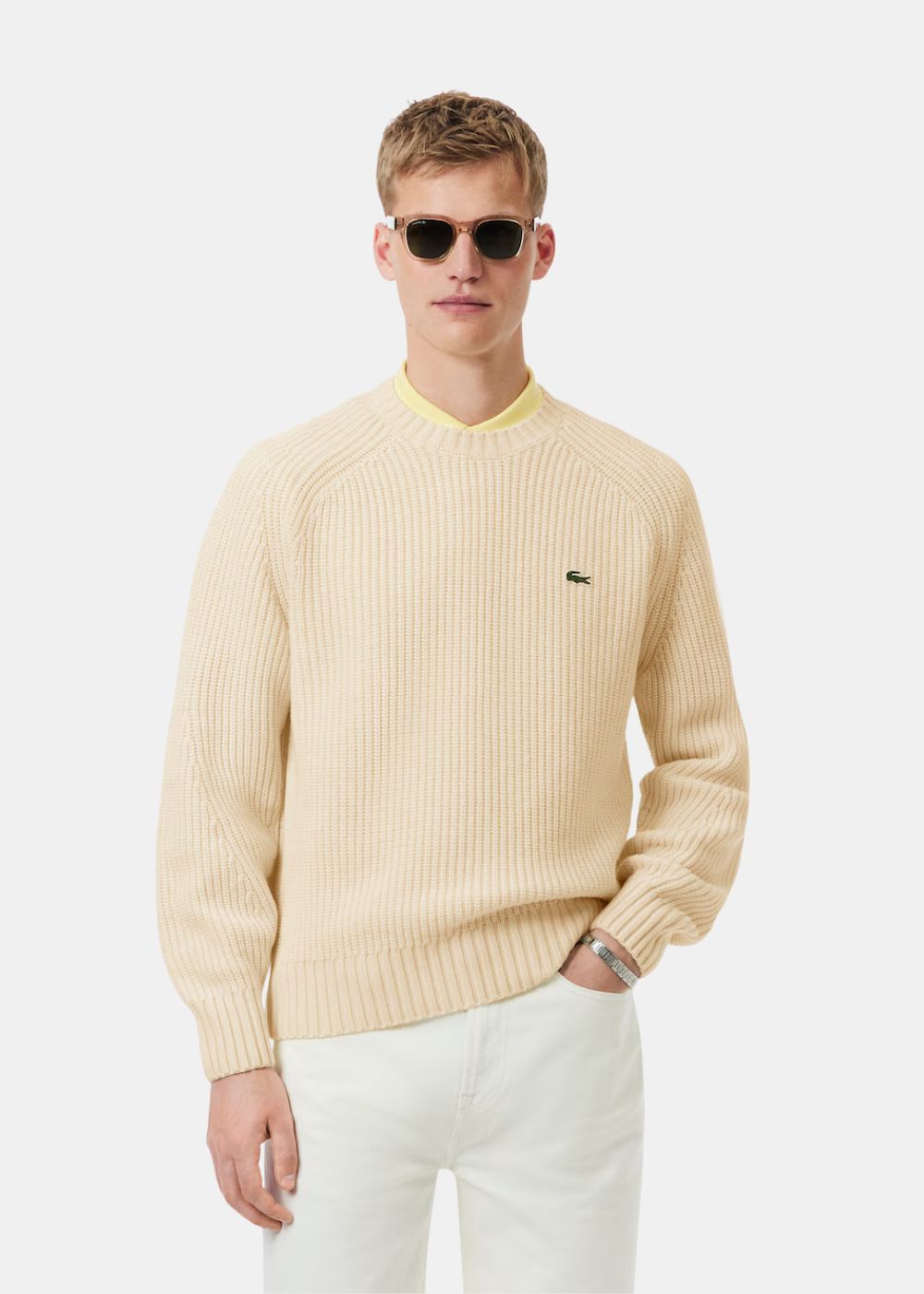 Mannequin portant le pull col rond Lacoste beige avec lunettes de soleil et pantalon blanc.