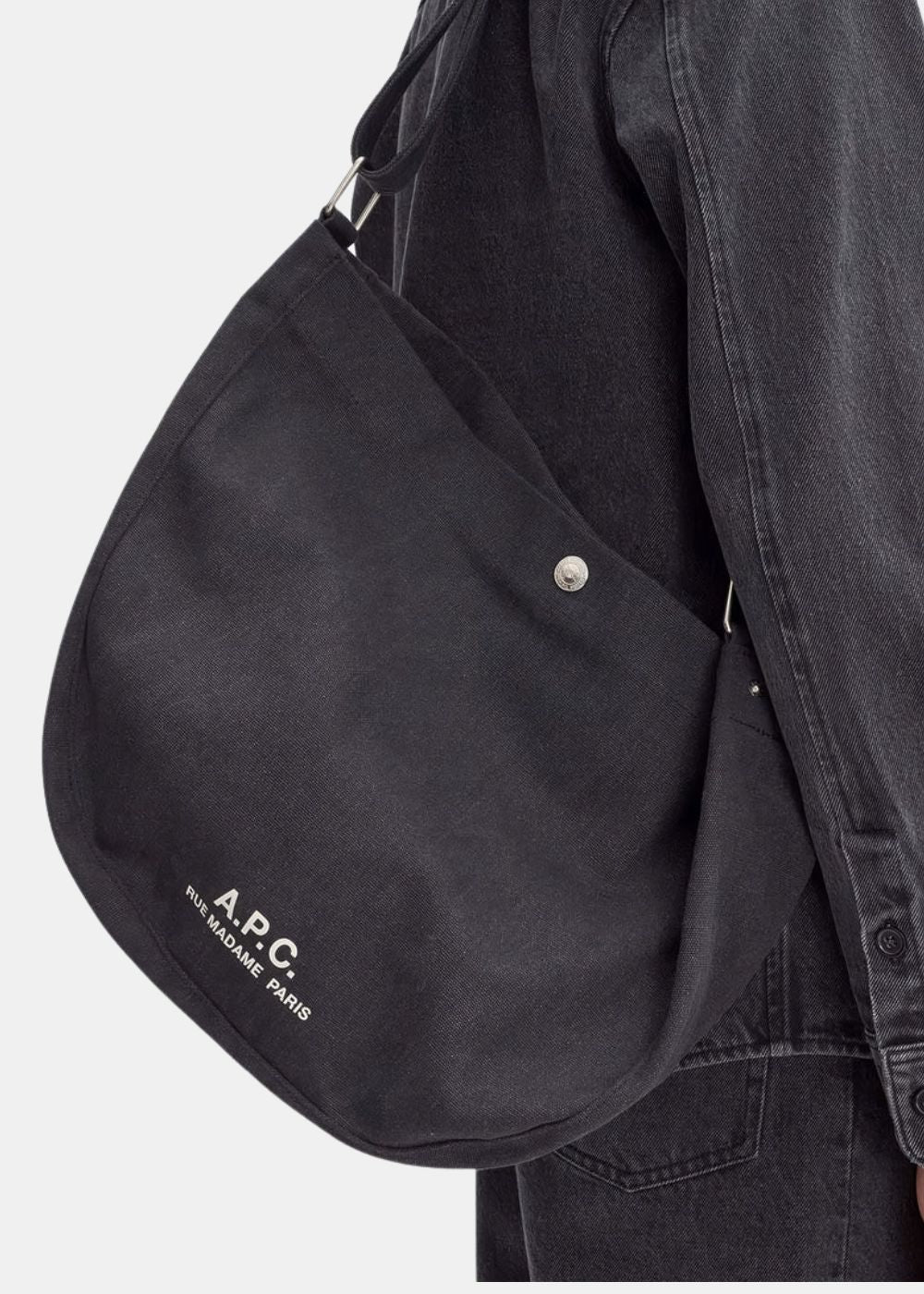 Zoom sur le porté épaule du sac besace A.P.C. noir. Permet de visualiser l'échelle du produit en situation, la texture brute de la toile de coton et le détail précis du logo blanc imprimé ainsi que du bouton pression métallique.