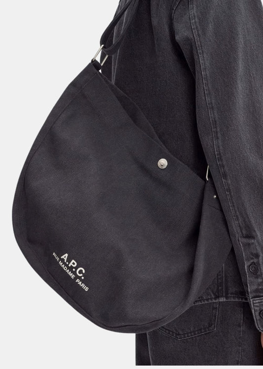 Zoom sur le porté épaule du sac besace A.P.C. noir. Permet de visualiser l'échelle du produit en situation, la texture brute de la toile de coton et le détail précis du logo blanc imprimé ainsi que du bouton pression métallique.
