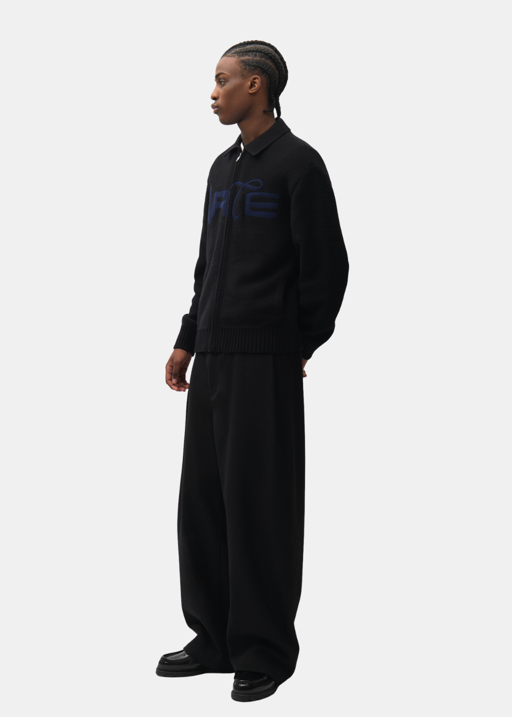 Mannequin de trois-quarts portant le cardigan zippé noir Arte-Antwerp avec un pantalon noir ample et des chaussures vernies.