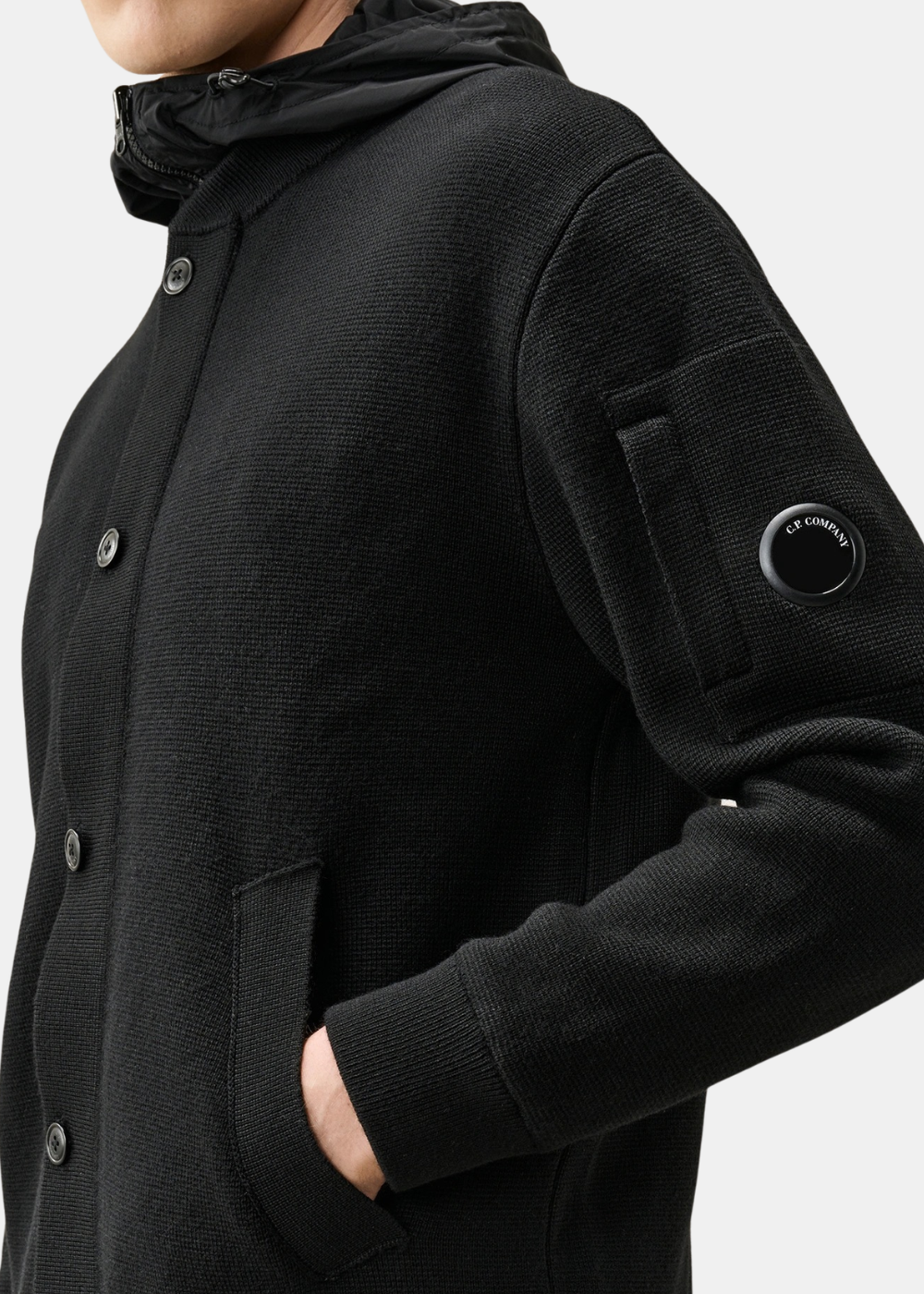 Gros plan sur le côté maille en laine mérinos noire du cardigan C.P. Company avec fermeture boutonnée, poche passepoilée et poche sur la manche ornée de l'emblématique lentille noire