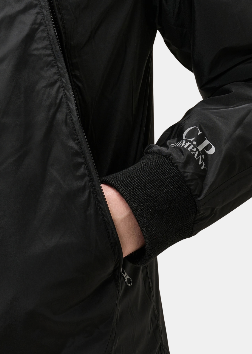 Gros plan sur la manche du cardigan réversible C.P. Company côté nylon noir, montrant le logo imprimé en blanc, le poignet côtelé et la poche zippée