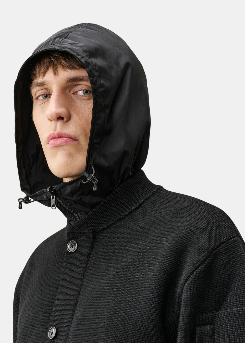 Gros plan sur le mannequin portant la capuche en nylon noir à cordons de serrage, contrastant avec le col en maille du cardigan