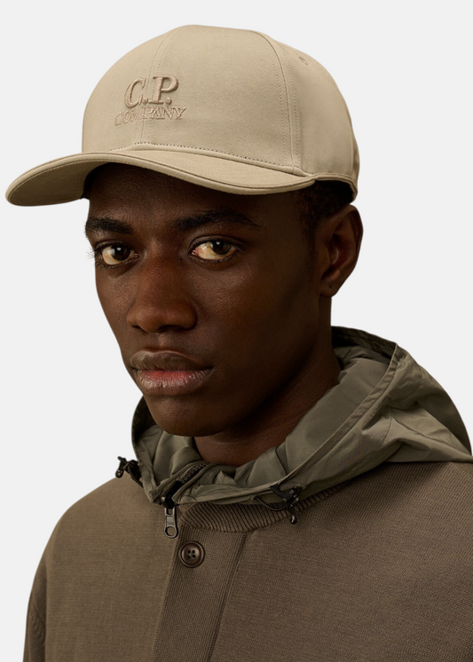 Gros plan sur le visage d'un mannequin noir de face portant la casquette C.P. Company beige sur fond uni blanc
