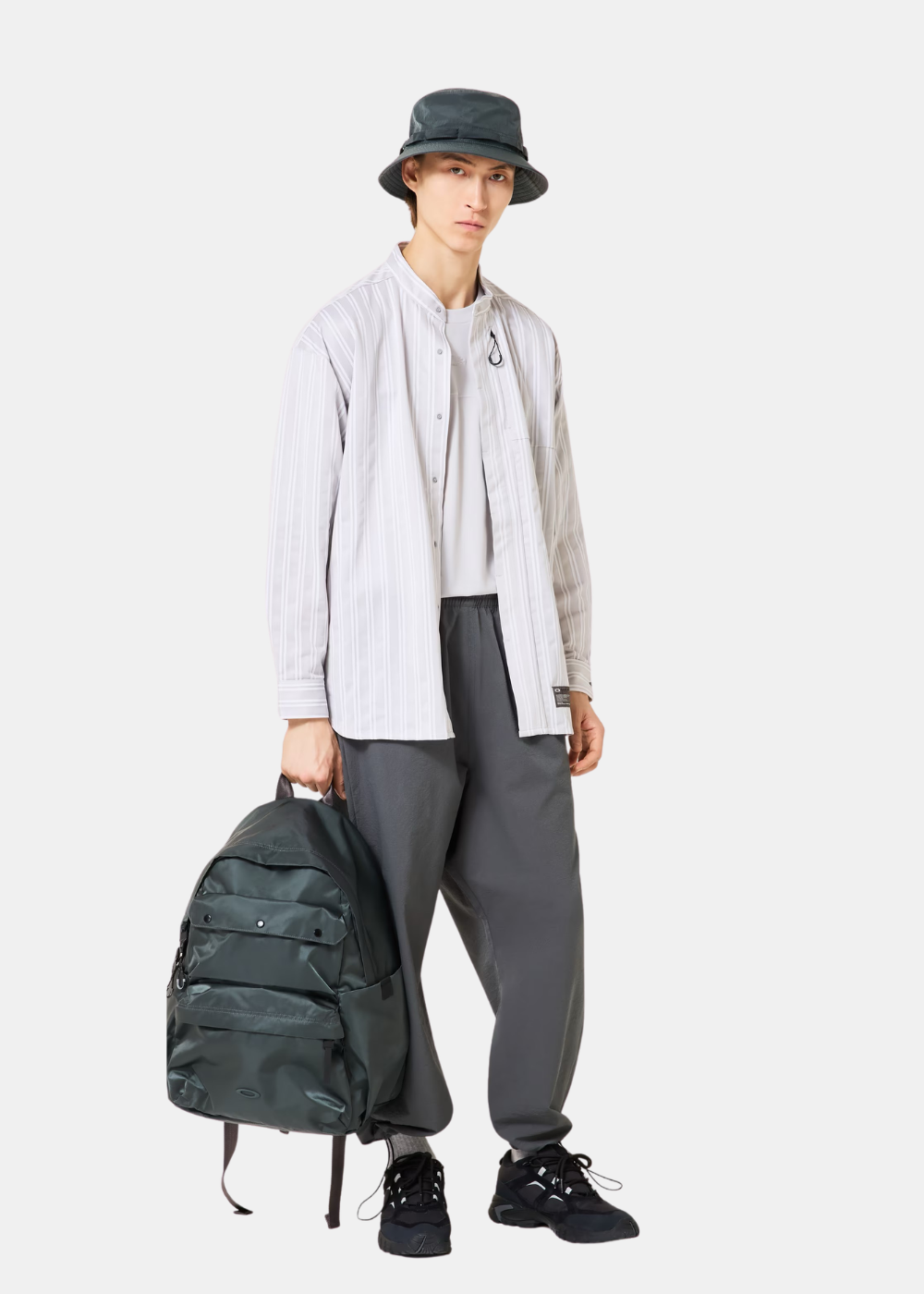 Mannequin portant la chemise Oakley blanche à rayures, un pantalon noir, un bonnet et un sac à dos, de face.