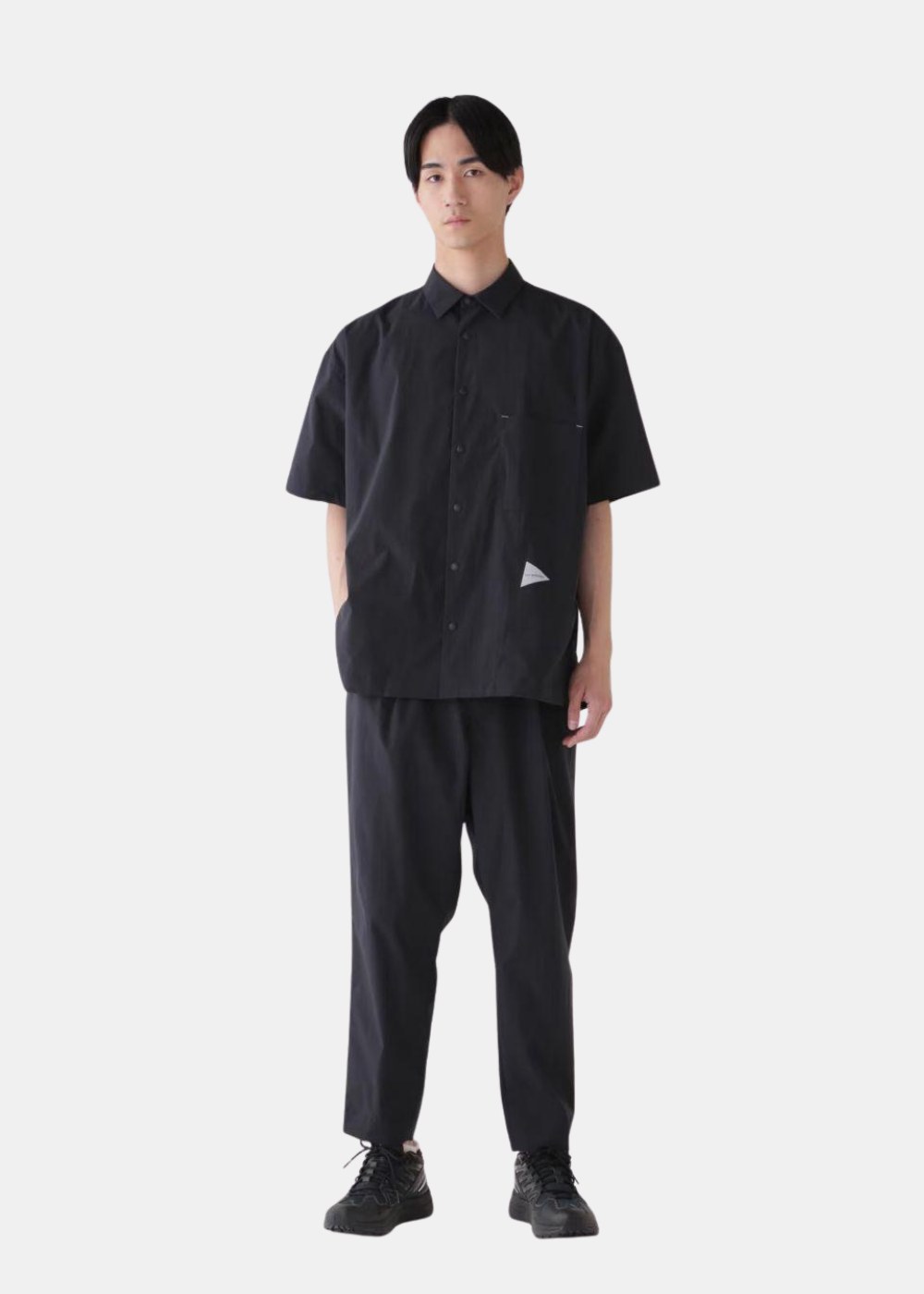 Look complet de face composé de la chemise noire And Wander, d'un pantalon fluide assorti et de baskets sombres.