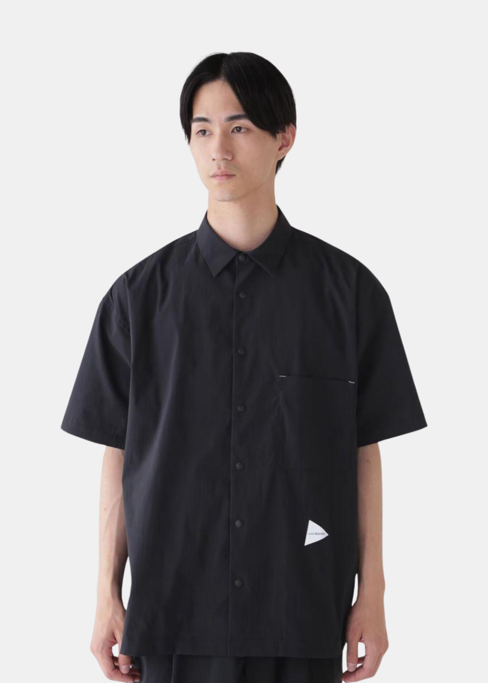 Chemise anti-UV noire And Wander à manches courtes avec poche poitrine et logo triangulaire blanc, portée de face.