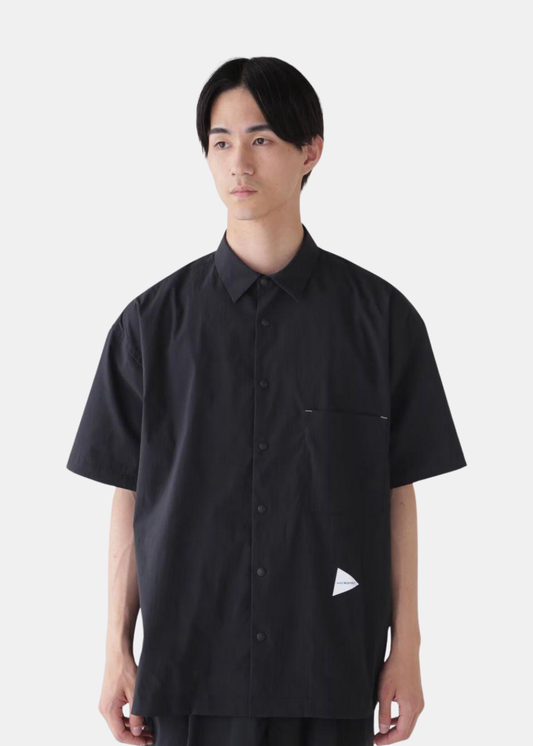 Chemise anti-UV noire And Wander à manches courtes avec poche poitrine et logo triangulaire blanc, portée de face.