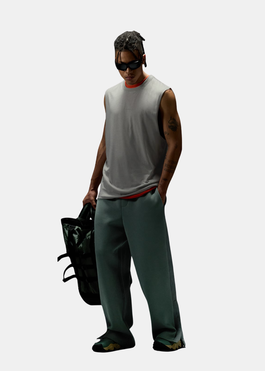 Un homme debout porte un débardeur gris Oakley, un pantalon large vert-bleu et tient un grand sac à dos vert foncé