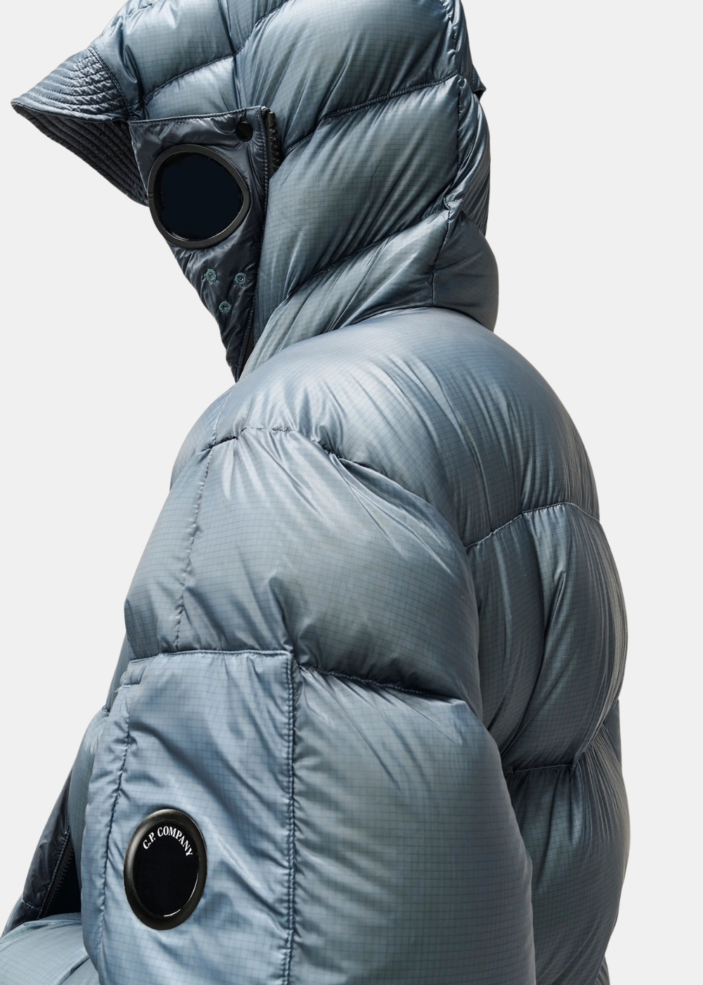Gros plan de profil sur la manche de la doudoune C.P. Company bleue montrant la poche avec la lentille signature et la capuche Goggle