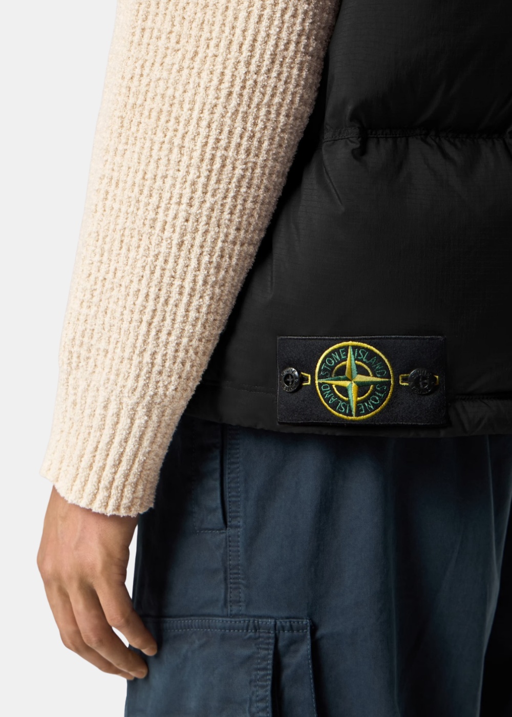 Gros plan sur le badge boussole Stone Island bouttonné sur le bas côté gauche de la doudoune sans manches noire