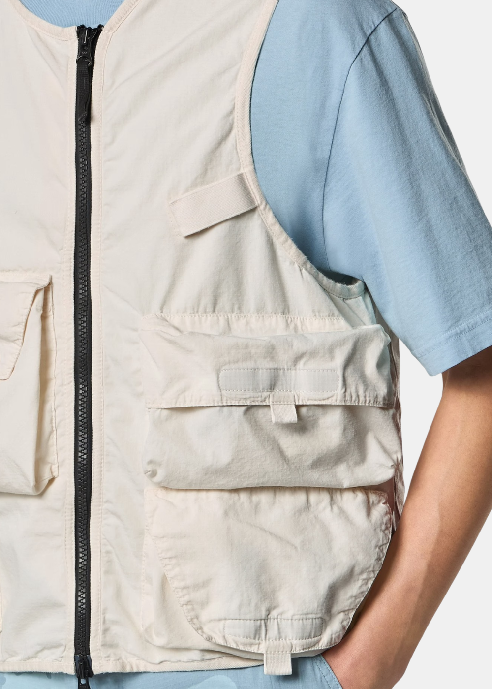 Gros plan porté du gilet Stone Island blanc en ripstop sur t-shirt bleu ciel, focus sur le zip central et les poches utilitaires
