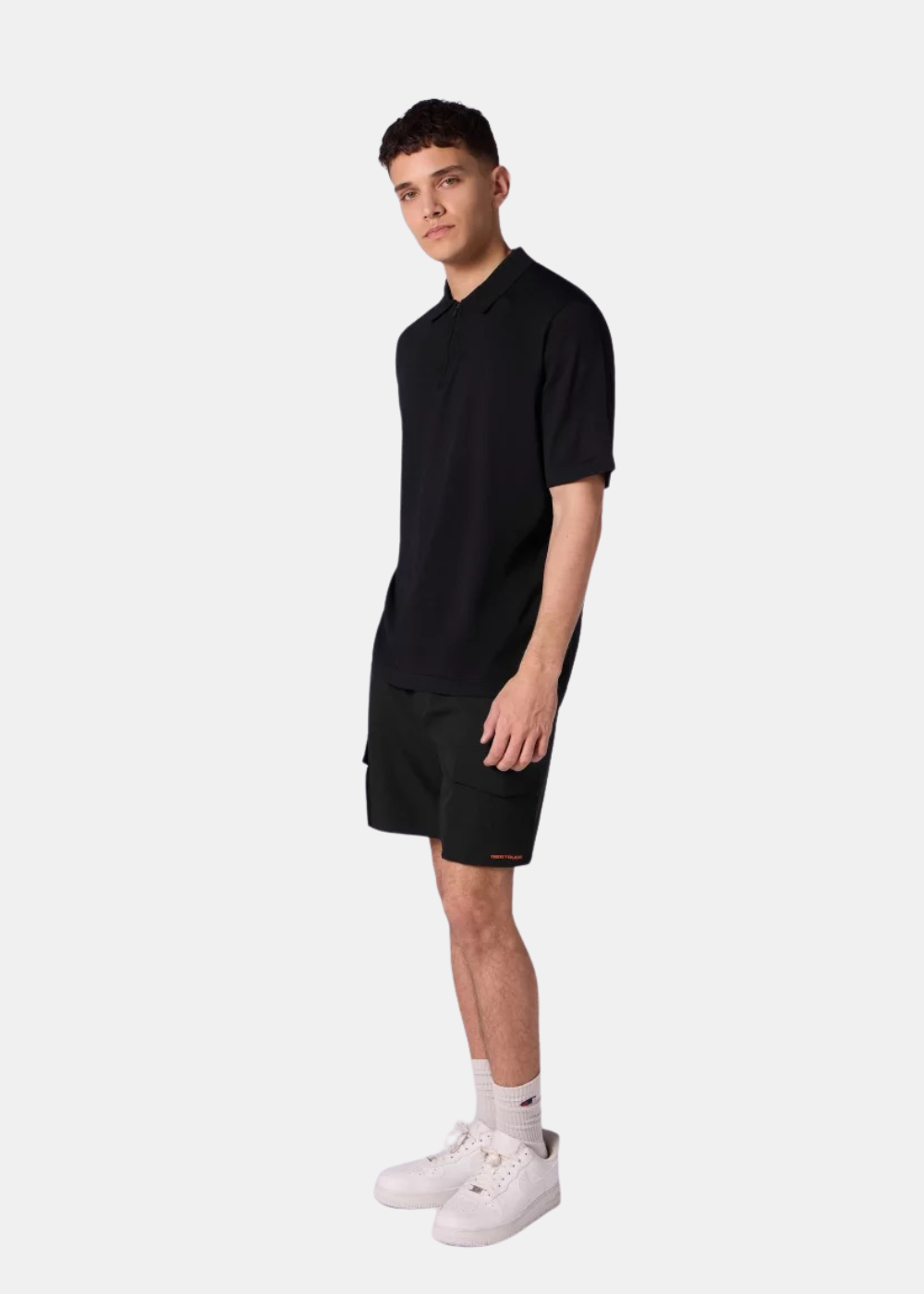 Un mannequin homme porte le polo oversize en maille noir Gertrude Jude avec un short assorti en maille noir au-dessus des genoux.
