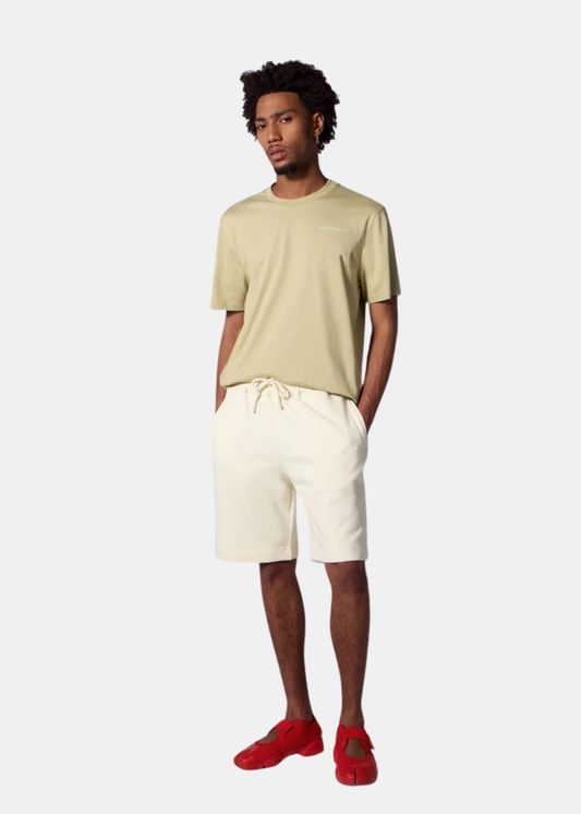 Un homme porte le short en molleton crème Gertrude Yann avec un t-shirt vert olive et des baskets rouges.