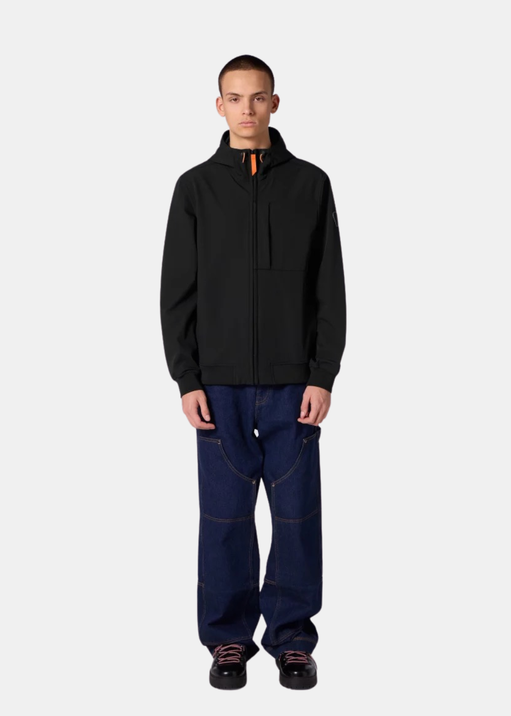 Un mannequin homme porte la veste technique Ronaldo noire Gertrude avec un jean large bleu foncé et des chaussures noires.