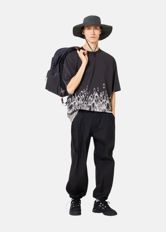 Un homme porte le pantalon technique noir Oakley avec un chapeau à larges bords, un t-shirt imprimé et un sac à dos