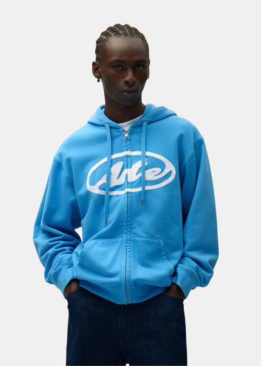 Hoodie zippé bleu Arte Antwerp avec logo blanc porté de face par un mannequin avec un pantalon jeans brut bleu. 