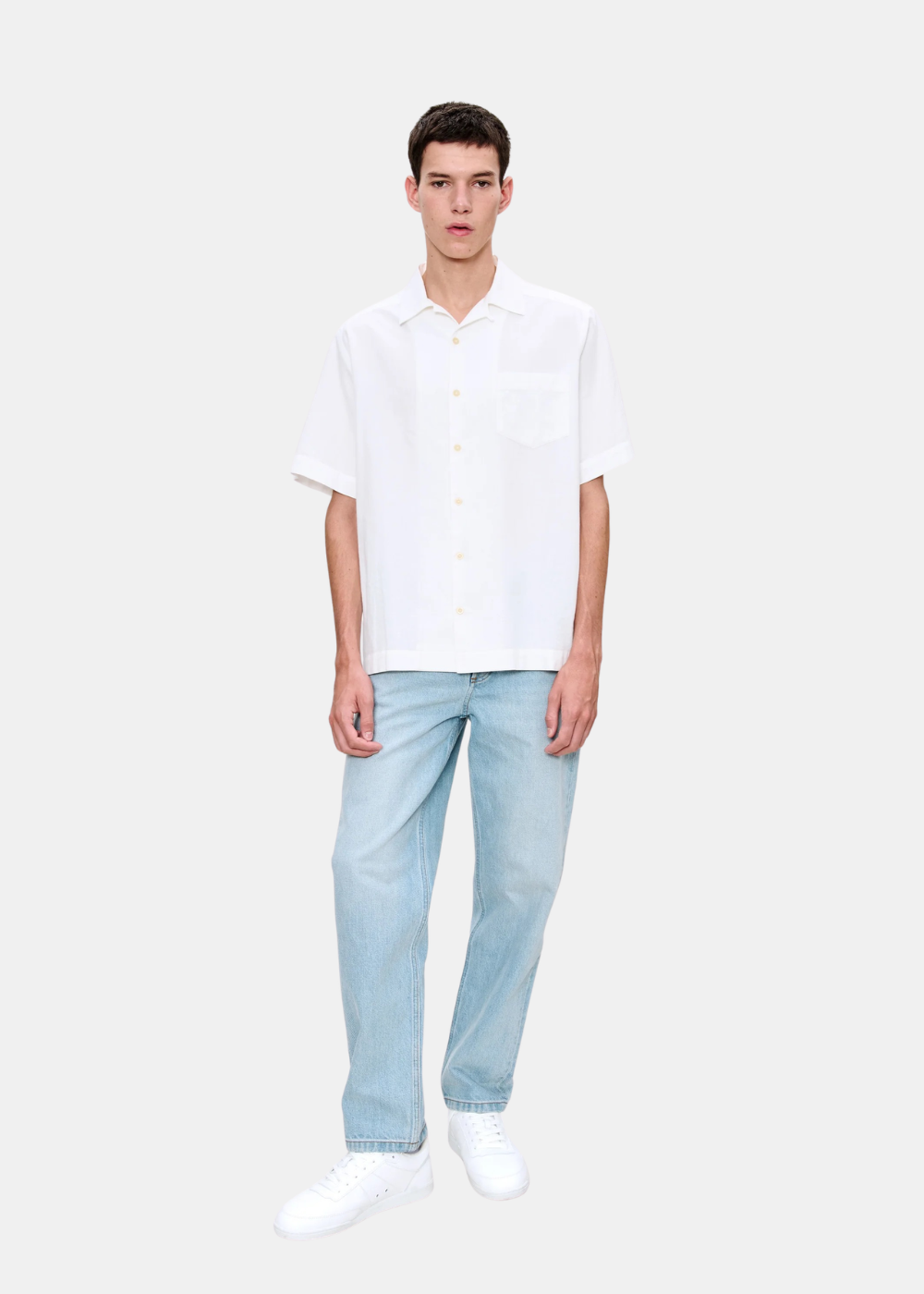 Portrait en pied d'un mannequin portant le jean droit ample bleu clair A.P.C. avec une chemise blanche et des baskets