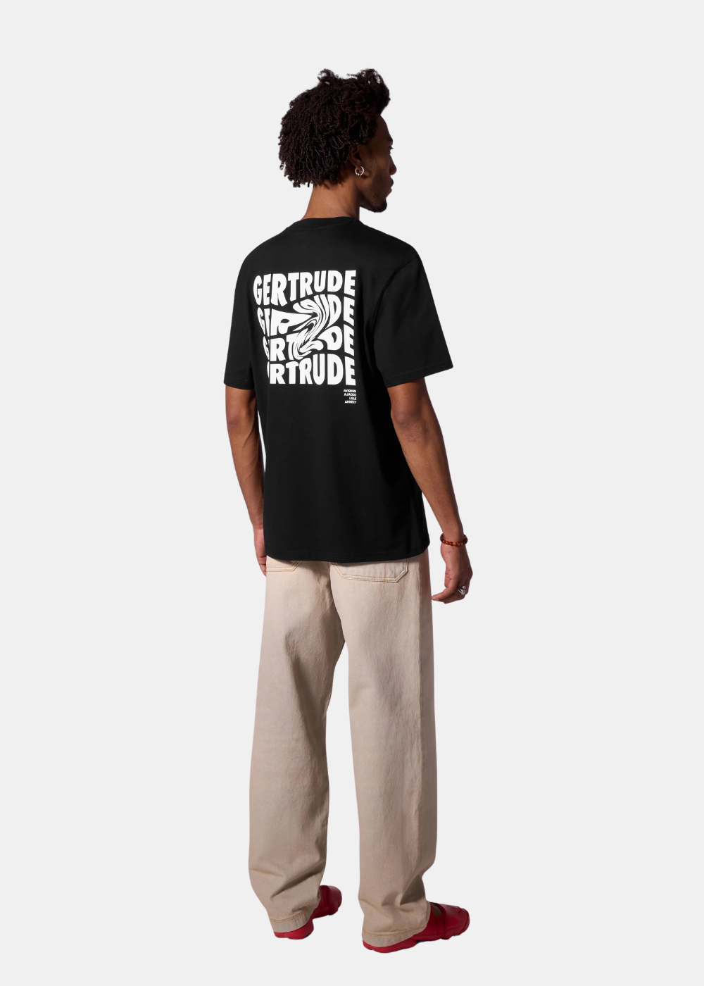Mannequin homme de dos portant le t-shirt noir Gertrude imprimé, un jean large beige et des chaussures rouges, sur fond uni.