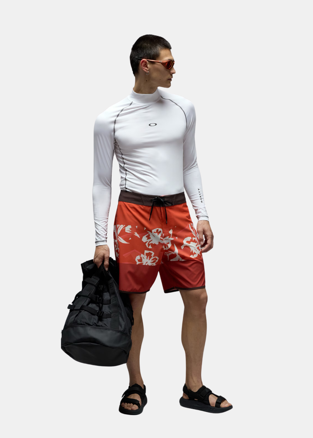 Homme portant un rashguard blanc, un short de bain rouge, des sandales noires et tenant un grand sac noir.