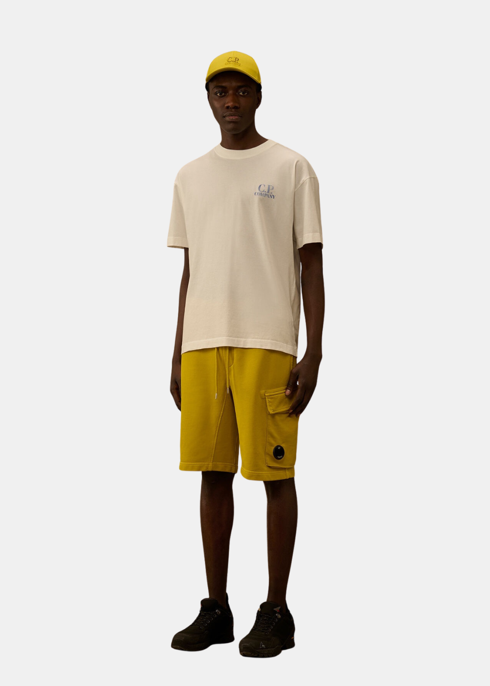 Mannequin portant le t-shirt blanc C.P. Company, un short cargo jaune et une casquette assortie