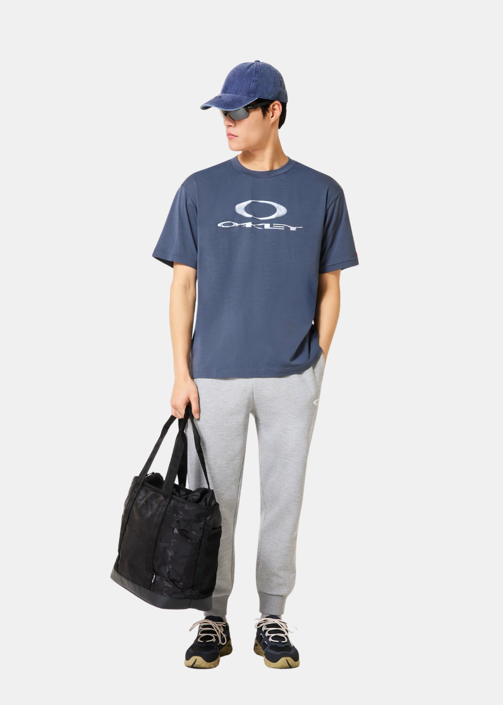 Homme portant un look complet : t-shirt bleu Oakley, jogging gris, casquette denim, lunettes de soleil et sac noir.