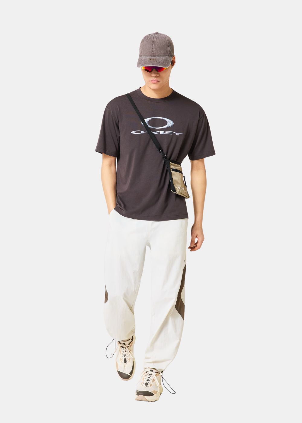 Homme portant le t-shirt gris Oakley, un pantalon blanc, une casquette grise et une petite sacoche beige.
