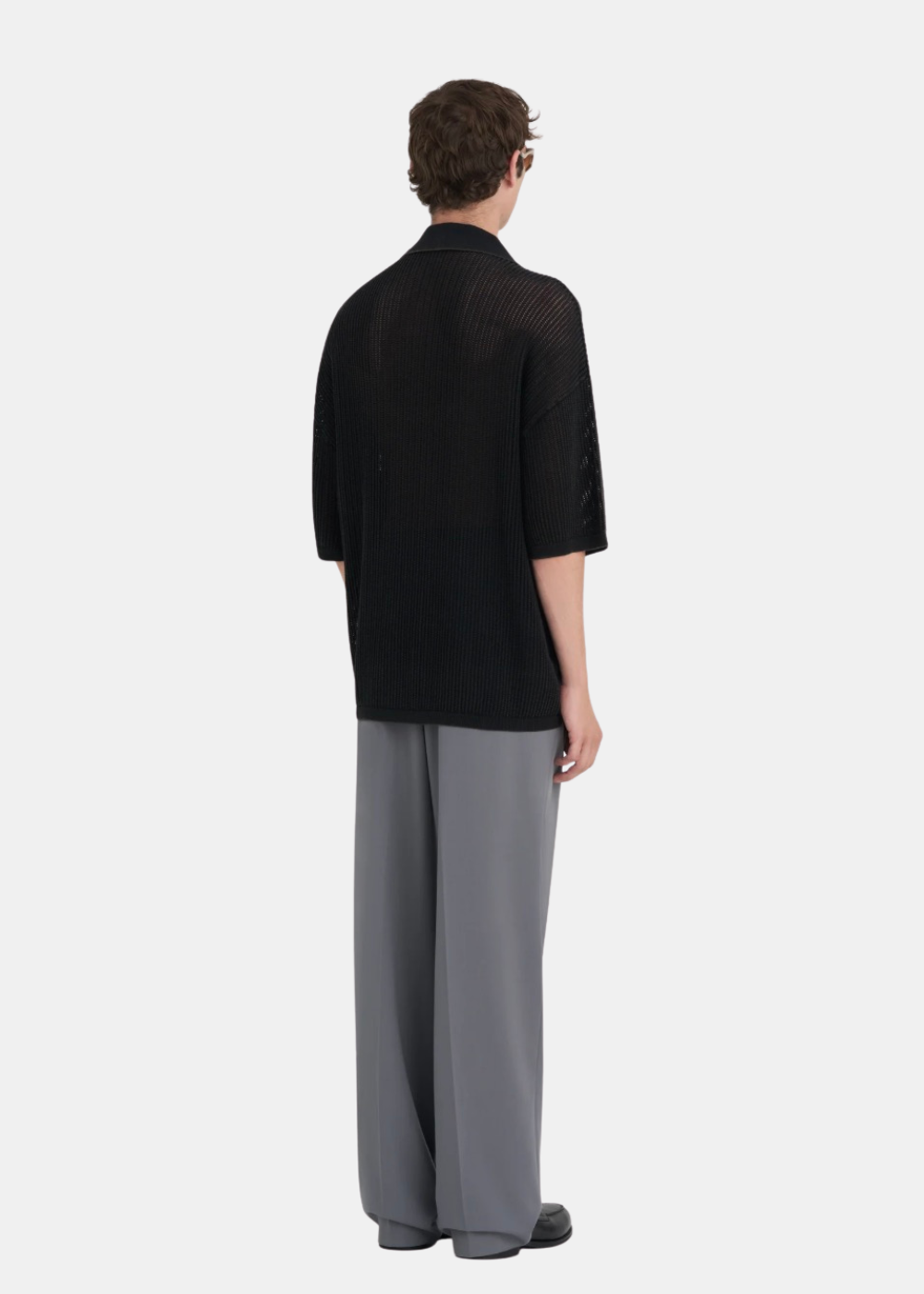 Homme portant le polo en maille noire Études Studio, un pantalon large gris et des lunettes de soleil Oakley.