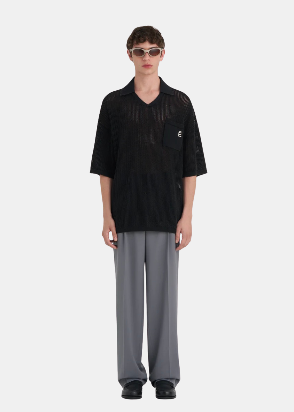 Homme portant le polo en maille noire Études Studio, un pantalon large gris et des lunettes de soleil Oakley.