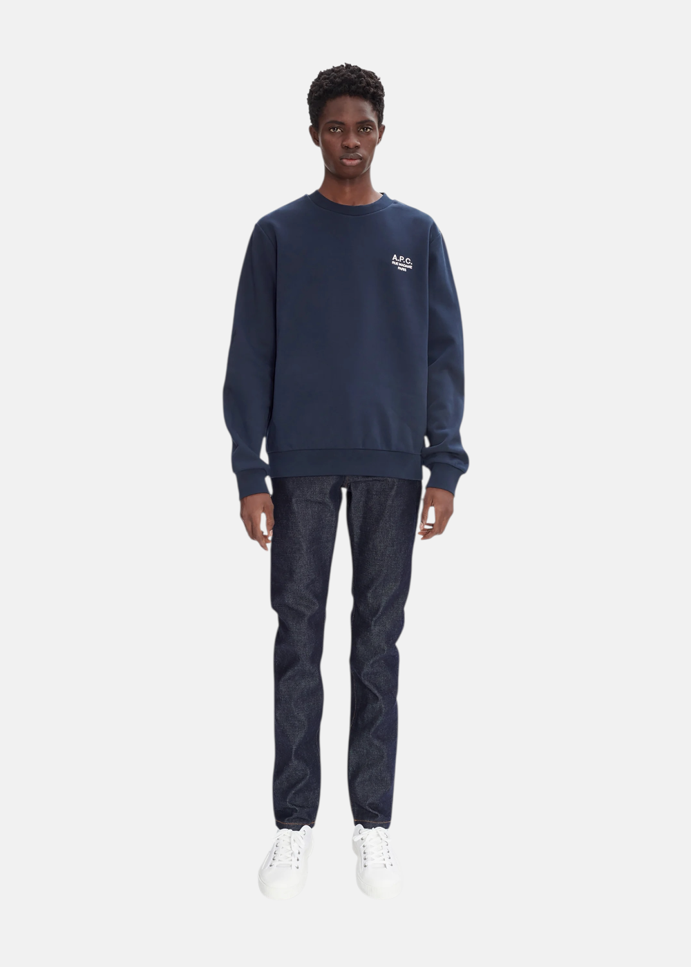 Homme portant le sweat A.P.C. bleu marine avec un jean brut bleu et des chaussures blanches, silhouette complète.