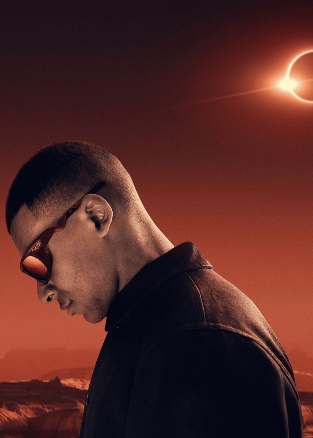 Portrait de profil en contre-plongée de Kylian Mbappé tête baissée, portant les lunettes Oakley Lateralis Prizm Ruby sur un fond rouge désertique surmonté d'une éclipse solaire lumineuse