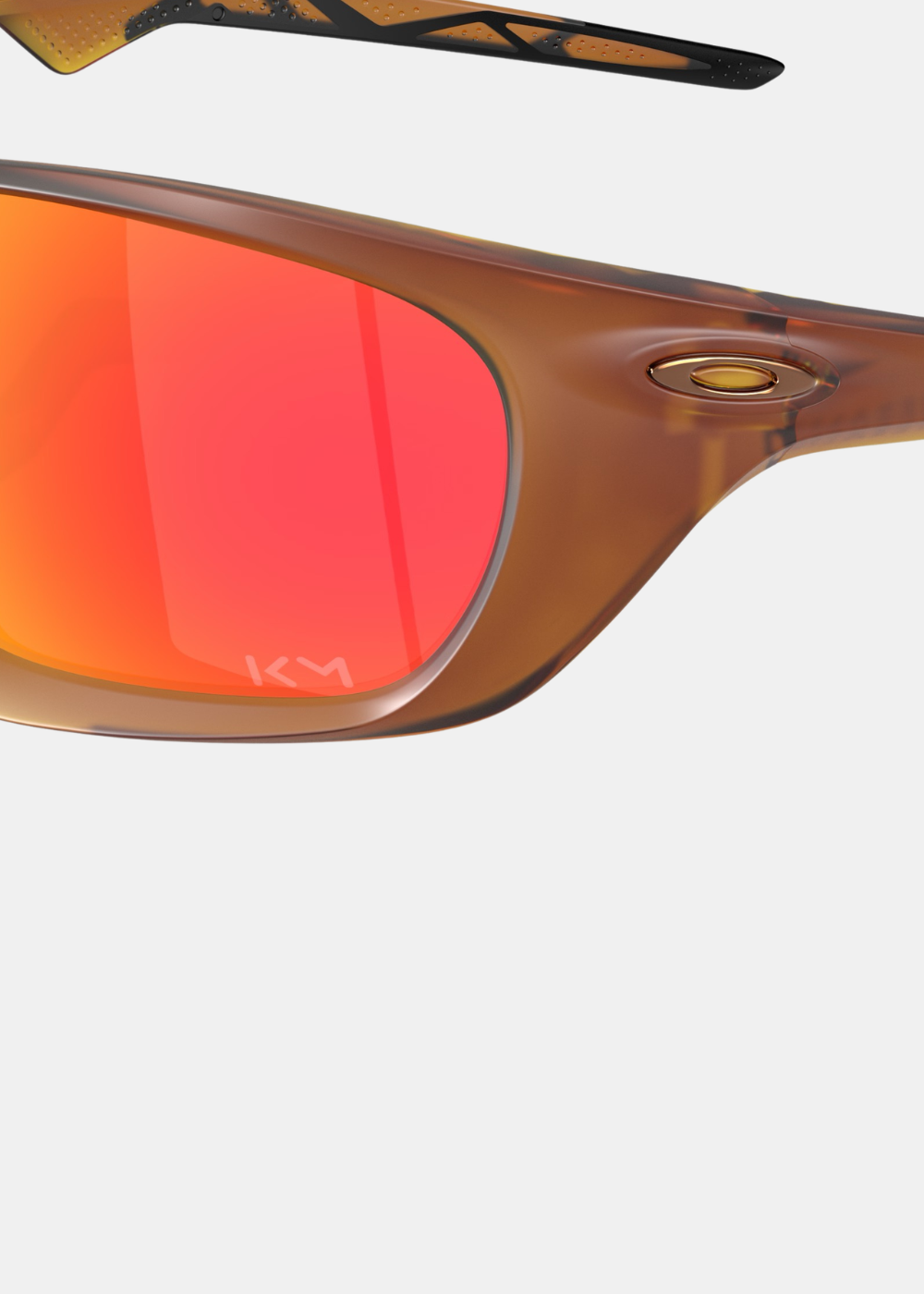 Gros plan détaillé sur le coin gauche des lunettes Oakley Lateralis Mbappé, affichant l'icône ovale dorée intégrée à la monture marron écaille et la gravure laser KM sur le bas du verre miroir Prizm Ruby
