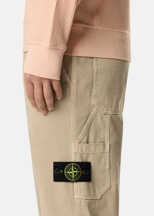 Gros plan sur le badge Stone Island amovible sur la poche latérale d'un pantalon beige en coton.