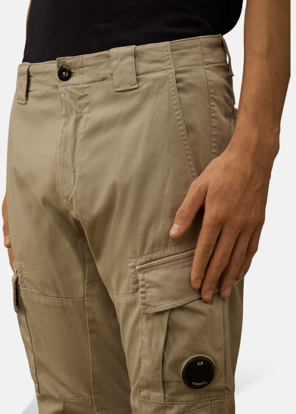 Gros plan sur la poche cargo du pantalon beige C.P. Company montrant le détail iconique de la lentille noire