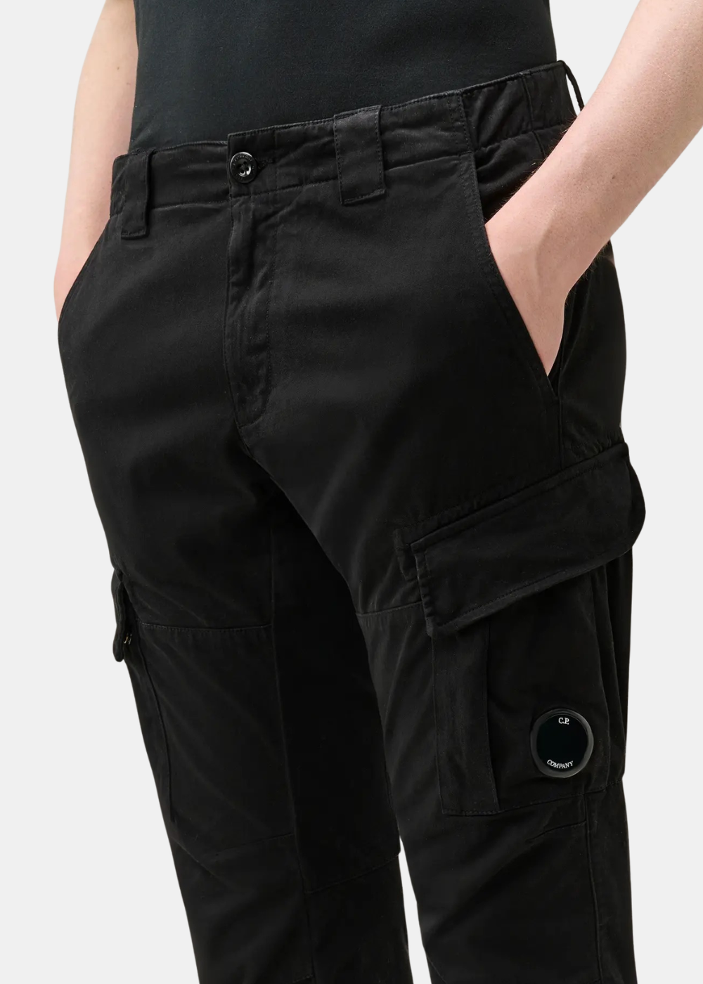 Gros plan de face sur un pantalon cargo noir CP Company en coton stretch avec les mains dans les poches et le verre de lunette signature