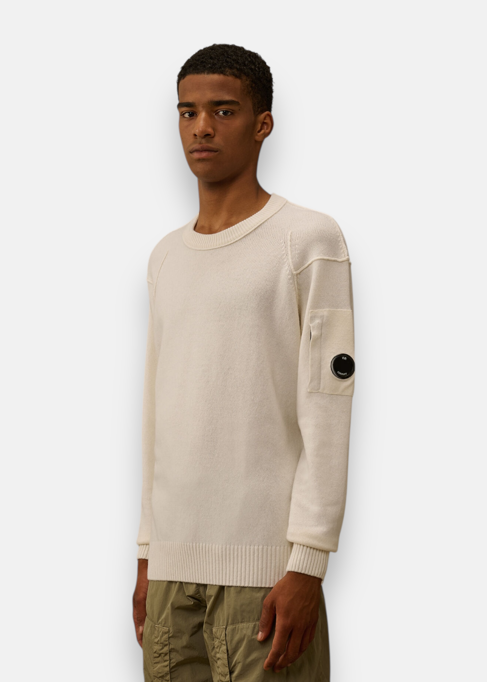 Homme portant le pull C.P. Company Lambswool col rond beige, vue de profil, montrant la lentille signature.