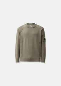 Packshot de face sur fond gris d'un pull col rond C.P. Company en laine lambswool GRS couleur kaki, avec détails de coutures apparentes aux épaules et poche à lentille sur la manche gauche