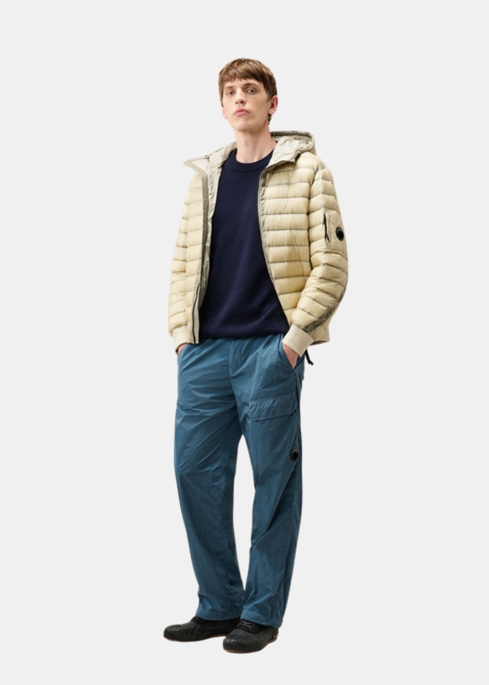 Porté en pied d'un pull marine C.P. Company sous une doudoune légère beige, avec un pantalon bleu technique.