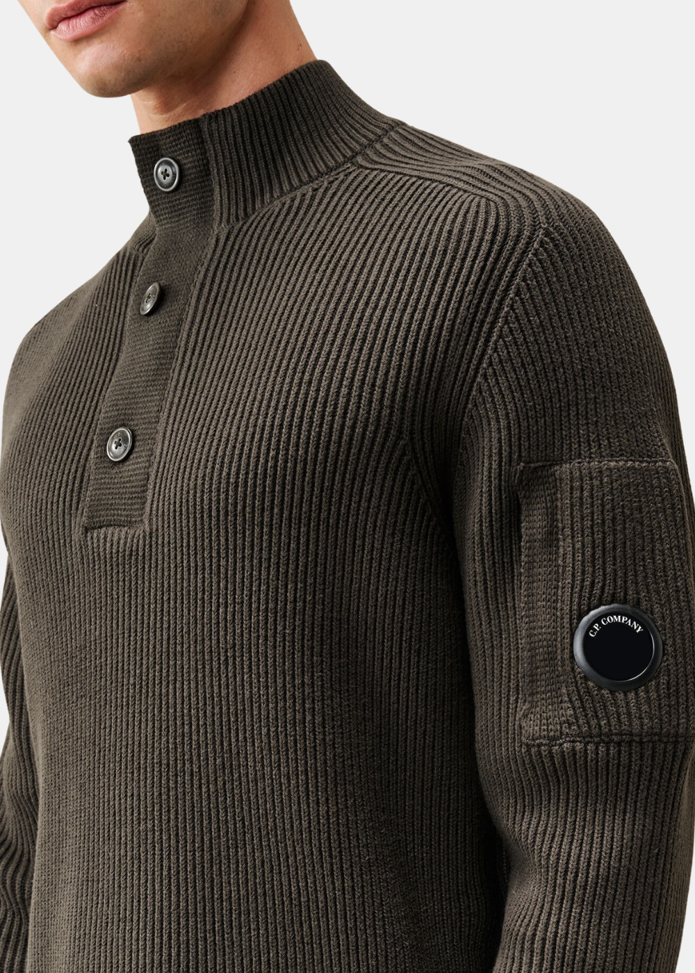 Gros plan sur le verre Lens C.P. Company et la pochette sur la manche gauche du pull polo gris