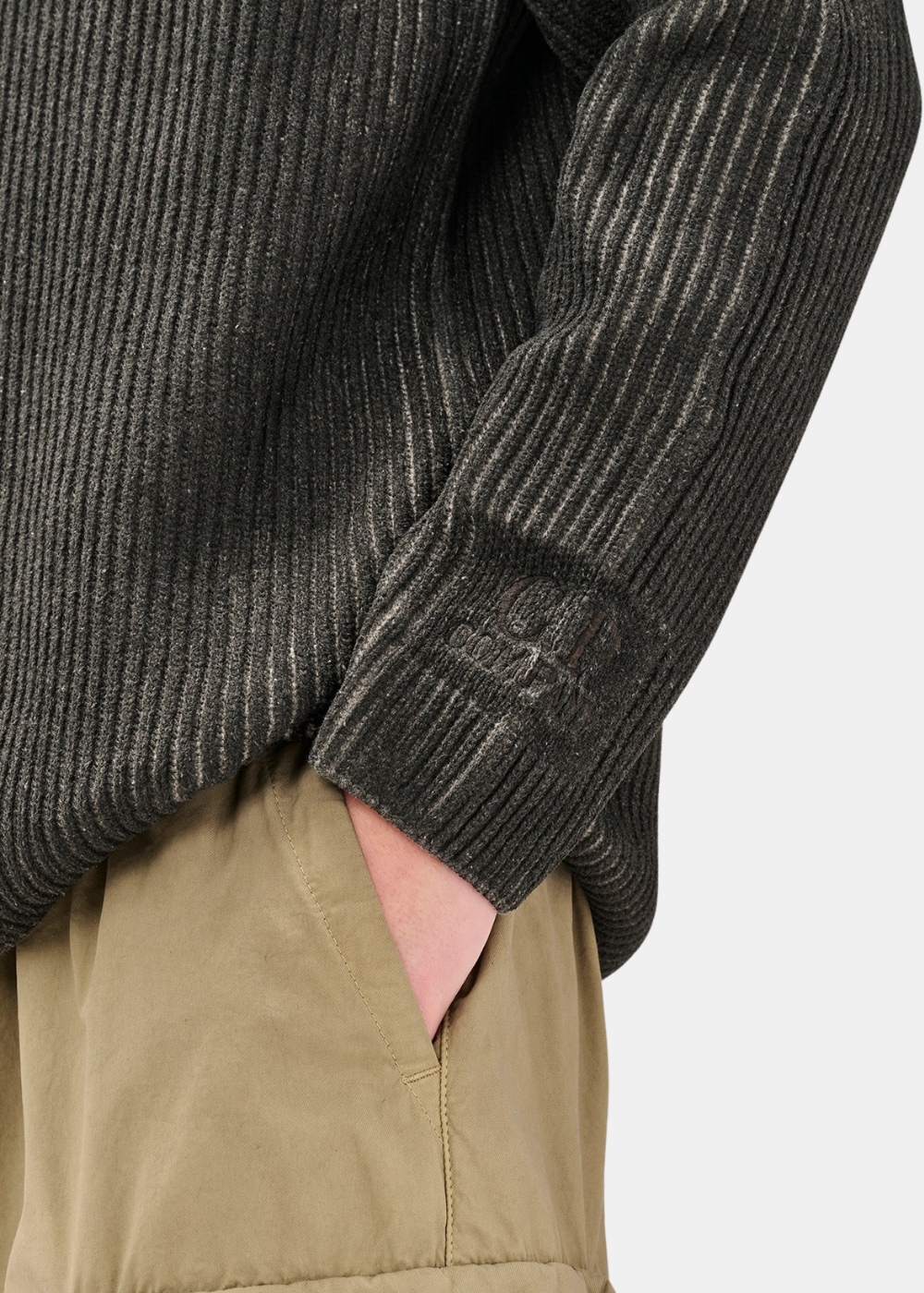 Gros plan sur le logo C.P. Company brodé ton sur ton sur le poignet gauche du pull à maille côtelée gris