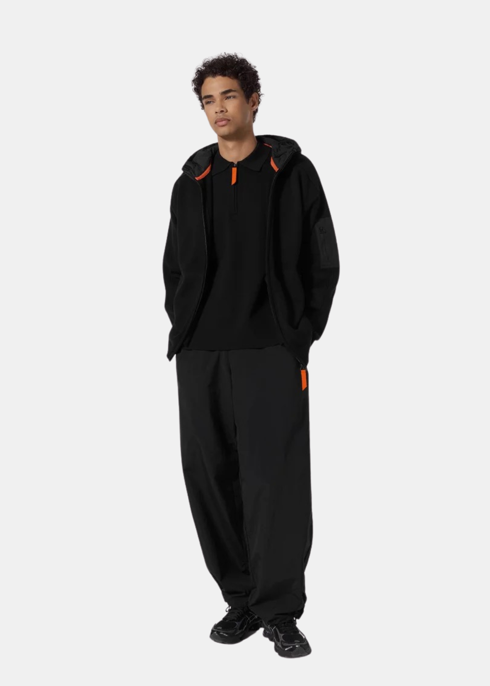 Mannequin homme de face en pied portant le pull zippé à capuche noir Gertrude ouvert sur un polo noir, un pantalon noir ample et des baskets noires
