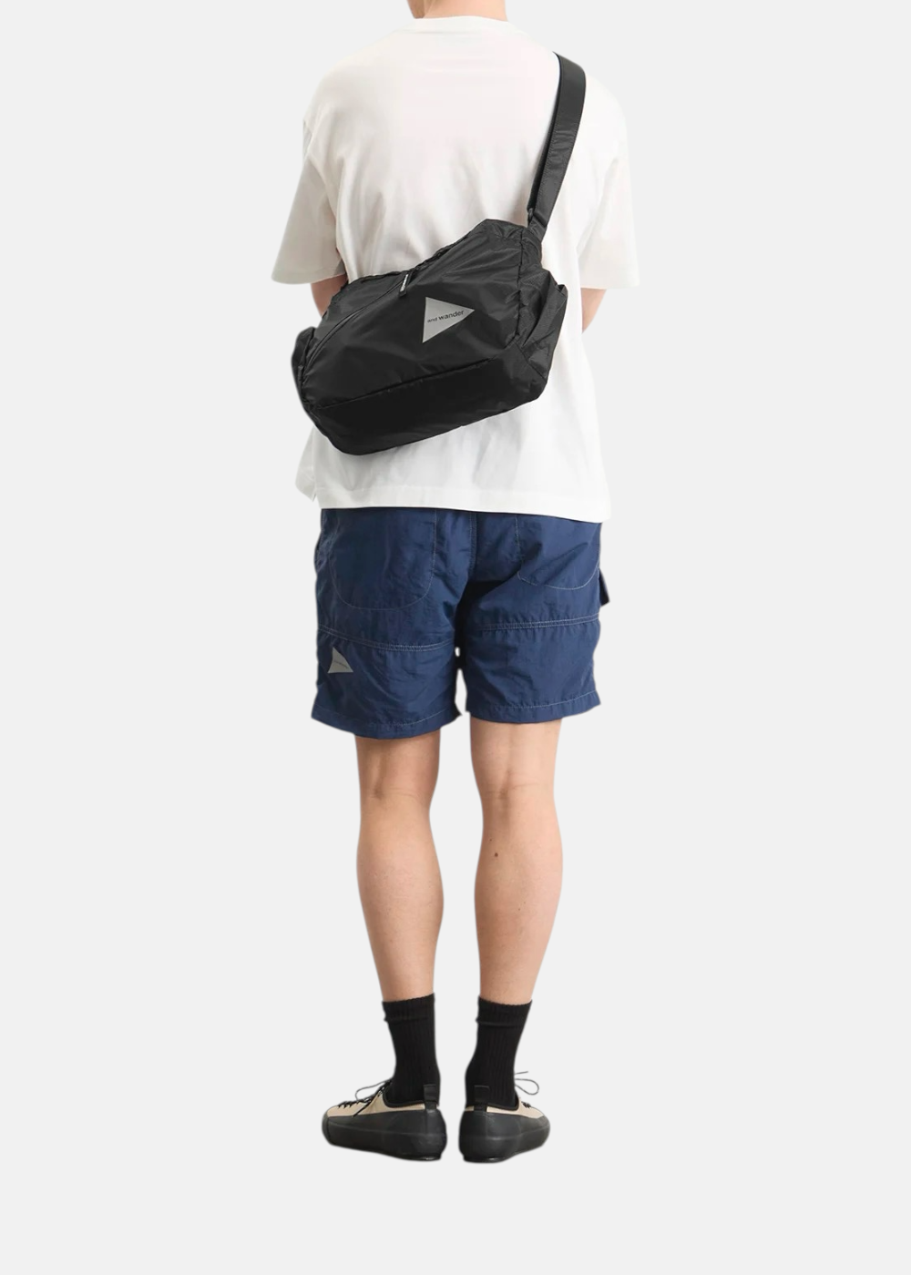 Sac bandoulière And Wander noir porté en travers du dos d'un homme en t-shirt blanc et short bleu