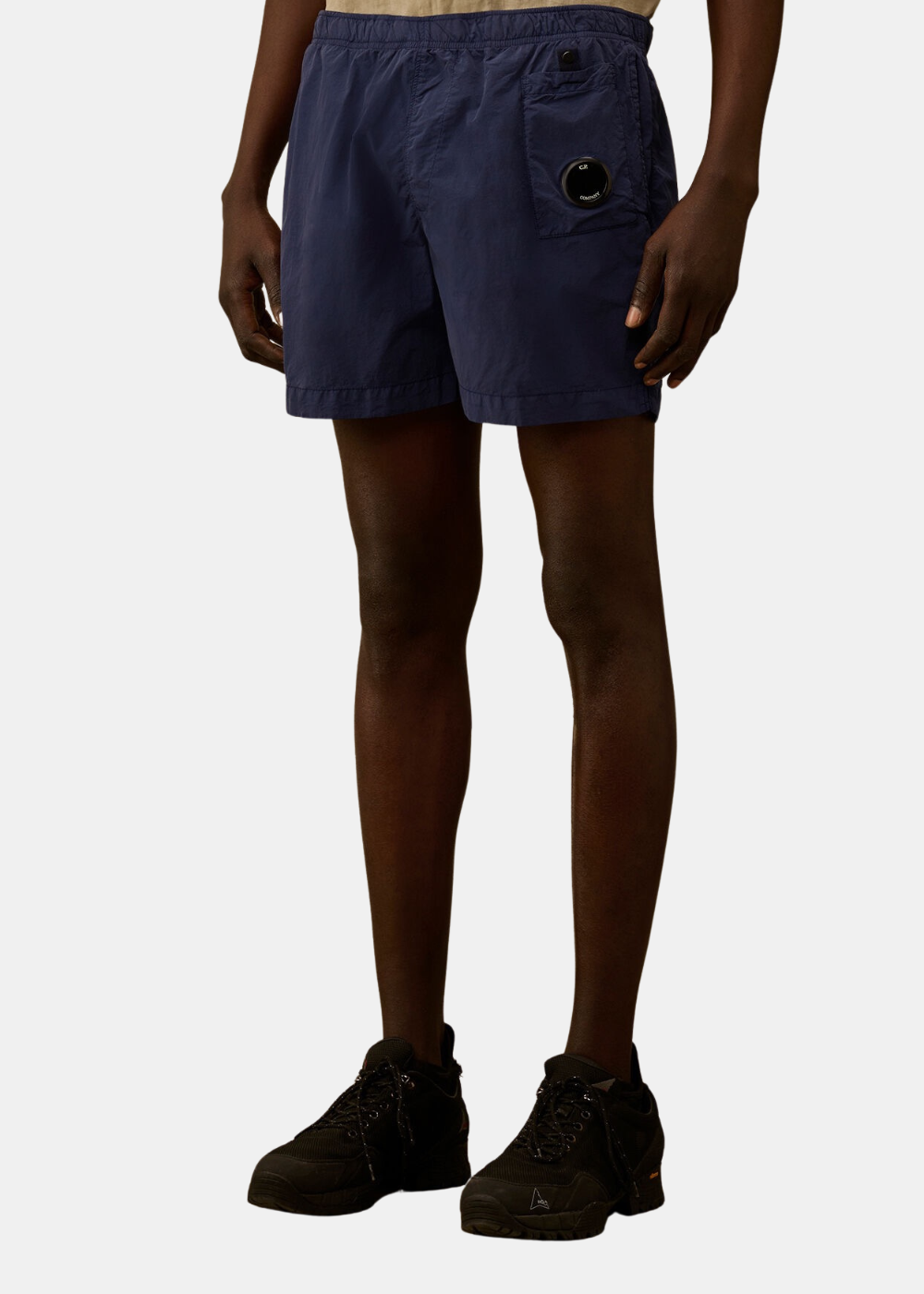 Mannequin homme de face portant le short de bain C.P. Company en nylon Flatt bleu marine, doté d'une taille élastique et d'une poche cargo plaquée sur la cuisse gauche avec l'emblématique lentille noire