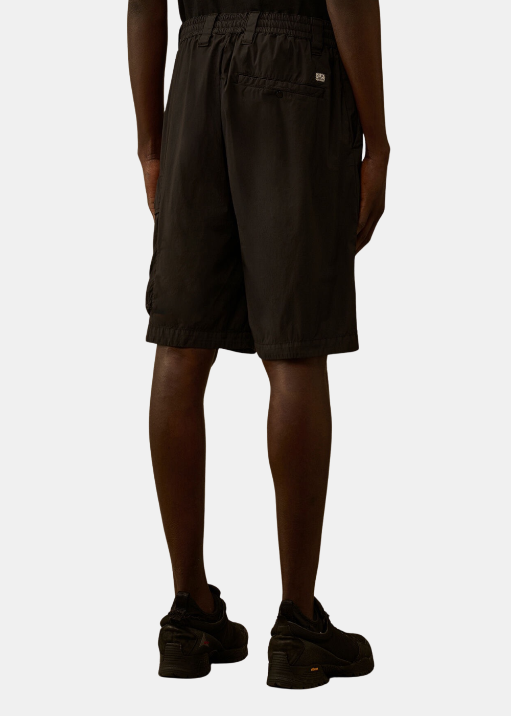 Mannequin homme vu de dos portant le short cargo C.P. Company noir en tissu 50 Fili stretch, montrant la coupe loose, la taille élastique avec passants de ceinture et une poche arrière boutonnée surmontée du petit écusson logo