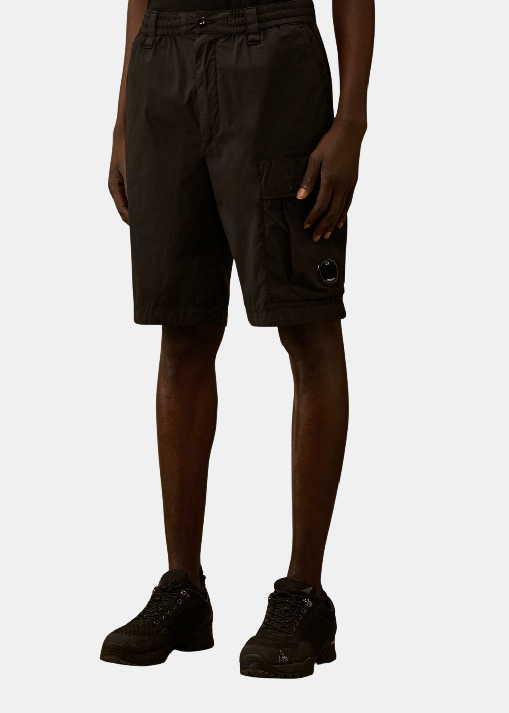 Mannequin homme de face portant le short cargo noir C.P. Company, mettant en valeur la coupe ample arrivant aux genoux et la grande poche cargo à rabat sur la cuisse gauche ornée de la lentille noire