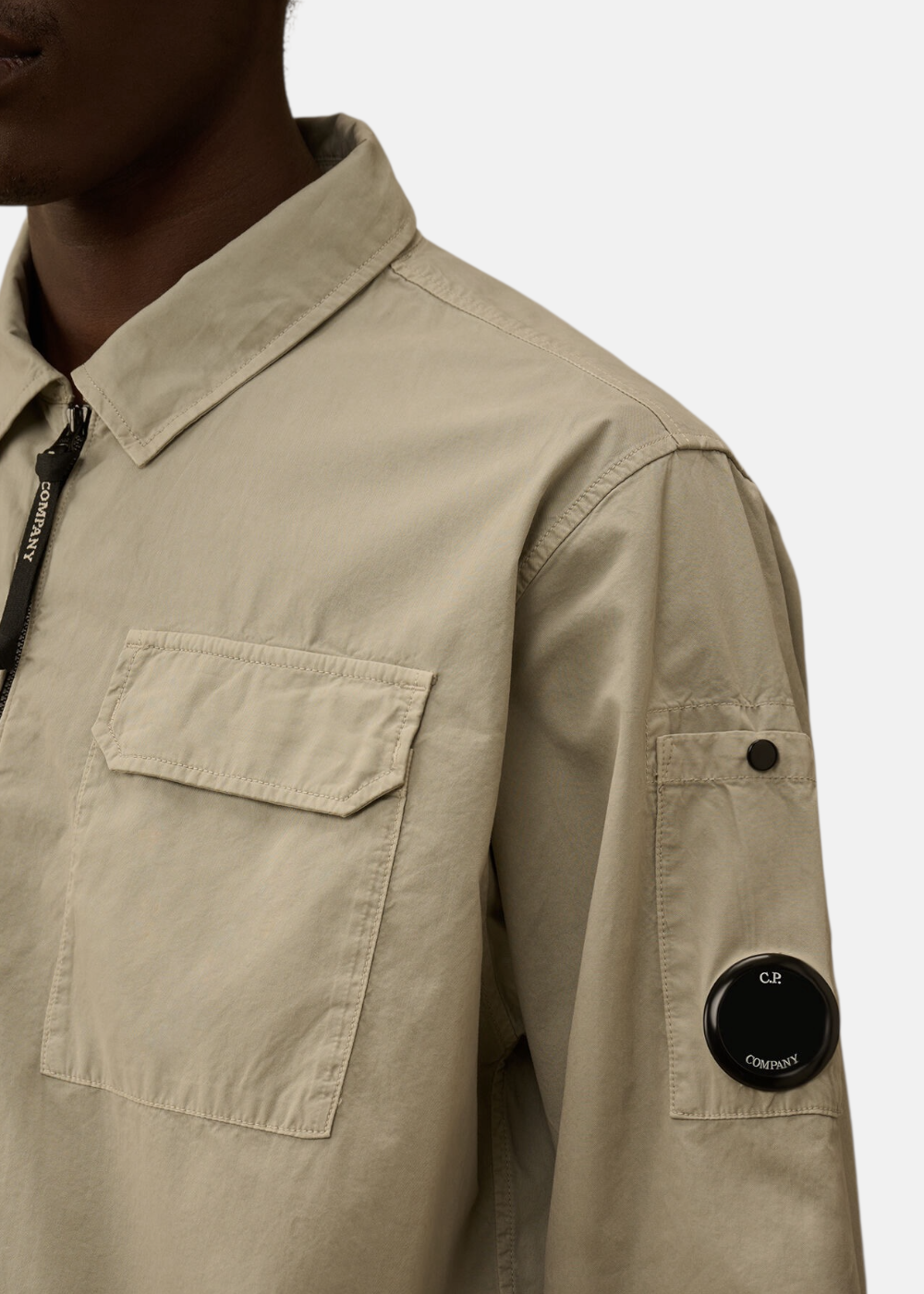 Gros plan sur le détail Lens noir sur la manche et le zip de la surchemise C.P. Company  beige.