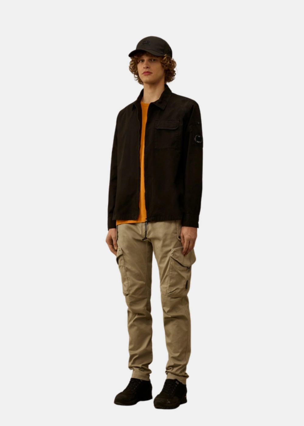 Porté en pied d'une surchemise noire C.P. Company ouverte sur un pull orange, avec pantalon cargo beige, casquette et chaussures sombres.