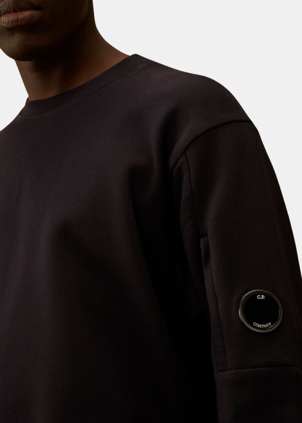Zoom sur la manche d'un sweat marine C.P. Company mettant en avant la lentille emblématique et la texture du tissu.