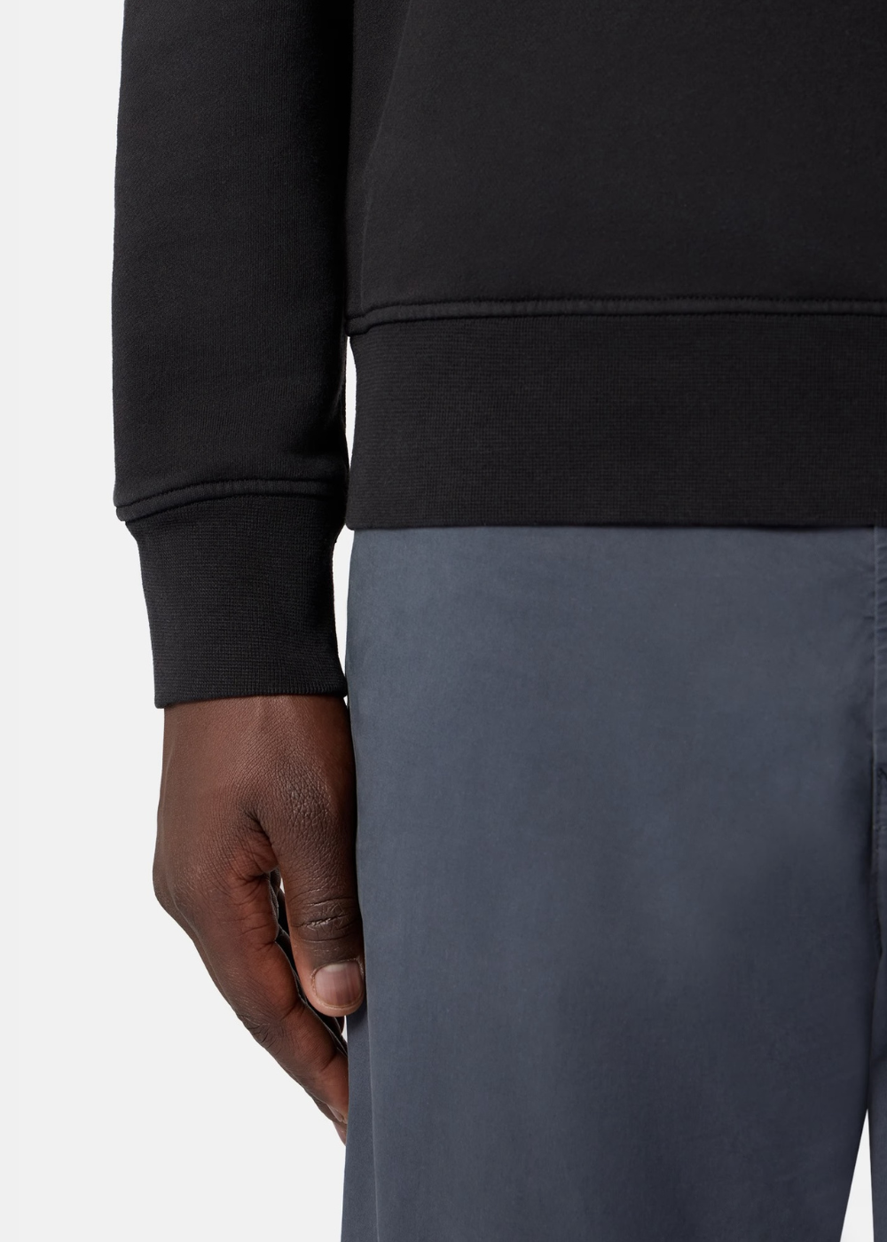 Zoom sur le bord-côte du poignet et de la taille d'un sweat noir Stone Island porté avec un pantalon bleu.